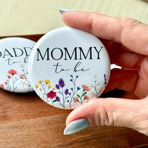 May include: White button with the text "Mommy to be" in black lettering with a floral design featuring colorful flowers and green stems.