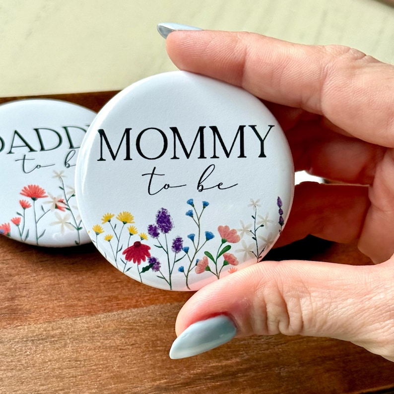 May include: White button with the text "Mommy to be" in black lettering with a floral design featuring colorful flowers and green stems.