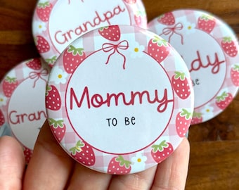 May include: A collection of round, pink and white buttons with strawberry and gingham patterns. The buttons have text that reads "Mommy to Be", "Grandpa", and "Granny". The buttons are decorated with strawberries and bows.
