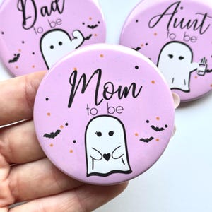 May include: Pink round buttons with the phrases "Mom to be", "Dad to be", and "Aunt to be" in black script. Each button features a white ghost illustration with black accents, bats, and orange dots.