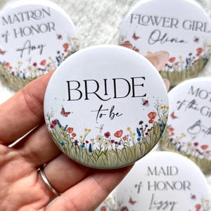 May include: White round buttons with floral designs and text for a wedding. The central button reads "BRIDE to be" with a wildflower border. Other buttons say "MATRON of HONOR", "FLOWER GIRL", "MOTHER of the GROOM", and "MAID of HONOR".