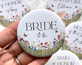 May include: White round buttons with floral designs and text for a wedding. The central button reads "BRIDE to be" with a wildflower border. Other buttons say "MATRON of HONOR", "FLOWER GIRL", "MOTHER of the GROOM", and "MAID of HONOR".