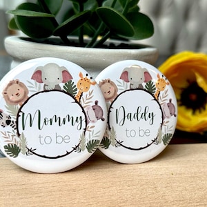Op de afbeelding: Twee witte ronde buttons met jungle dieren illustraties en de zinnen "Mommy to be" en "Daddy to be" in donkergroen schrift. De buttons tonen een olifant, een leeuw, een giraffe, een zebra en een nijlpaard.