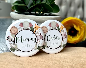 May include: Two white round buttons with jungle animal illustrations and the phrases "Mommy to be" and "Daddy to be" in a dark green script. The buttons feature an elephant, lion, giraffe, zebra, and hippo.