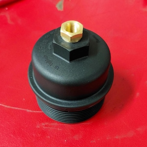 May include: Black oil filter housing with a gold-colored nut on top. The housing has a hexagonal shape and the text "25 Nm" and "to tighten" is visible. The filter is on a red surface.