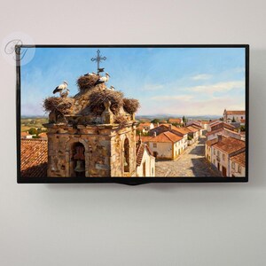 May include: A framed painting depicts a European village scene. The focal point is a stone bell tower topped with nests and storks. Red-tiled roofs and a blue sky complete the picturesque landscape. The artwork is displayed on a wall.