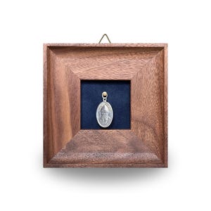 May include: A silver pendant depicting a religious figure, displayed within a wooden frame. The pendant hangs from a gold-coloured bail against a dark blue backdrop. The frame is a warm brown and has a hanging hook.