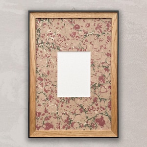 May include: A rectangular wooden picture frame with a black border. The frame holds a marbled paper design in shades of red, green, and gold on a tan background. A white rectangle is centered within the frame, creating a space for a photo or artwork.