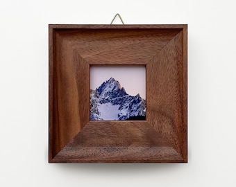 May include: Square wooden picture frame with a photograph of a snow-covered mountain peak. The frame is a warm brown color and has a simple, beveled design. A small metal hook is attached to the top for hanging.