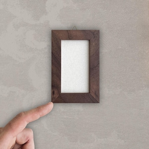 May include: A small, rectangular wooden picture frame with a dark brown finish. The frame has a white interior and is hanging on a light beige wall. A finger is pointing at the frame.