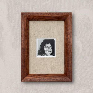 Peut inclure: Un portrait encadré en noir et blanc d'une personne posant sa main sur son visage. Le portrait est encadré dans un petit cadre carré, entouré d'un passe-partout en tissu beige et d'un cadre en bois brun foncé. L'œuvre est accrochée à un mur texturé blanc cassé.