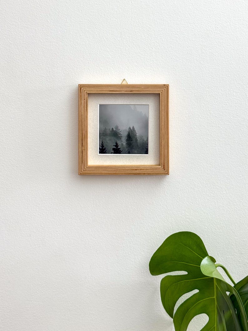 May include: Square framed art print featuring a misty forest scene. The artwork is displayed in a natural wood frame, hung against a white wall. The print showcases dark green trees shrouded in fog, creating a serene landscape.