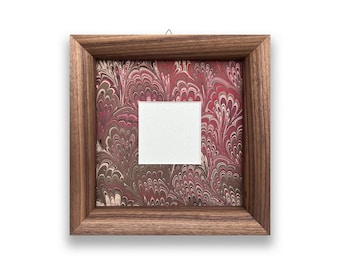 May include: Square wooden picture frame with a marbled red and white background. The frame has a square white opening in the center. The frame is hanging on a white background.
