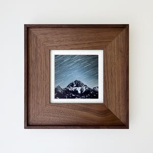 Puede incluir: Fotografía enmarcada de una cordillera bajo un cielo estrellado. La imagen está dentro de un marco de madera marrón oscuro. La fotografía captura los picos de las montañas y las rayas de luz de las estrellas.