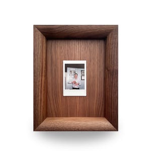 Peut inclure: Un coffret en bois brun foncé. À l'intérieur, une petite photographie est montée, montrant une personne tenant un objet. Le coffret a une étagère en bas. Le fond est blanc.