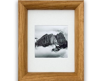 May include: A framed black and white photograph of a mountain peak shrouded in clouds. The photograph is set within a light brown wooden frame with a white mat. The frame is hanging on a white wall.