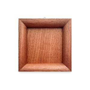 Puede incluir: Caja de sombra cuadrada de madera con un cálido acabado marrón rojizo. El marco tiene un borde biselado y un interior empotrado. Un pequeño colgador de metal está fijado en la parte superior para montaje en pared. La veta de la madera es visible.