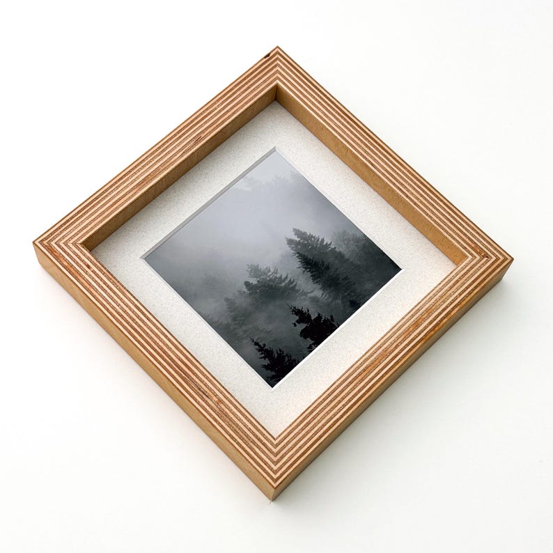 May include: Square wooden picture frame with a natural wood grain finish. The frame holds a black and white photograph of a misty forest scene. The frame has a white mat and is set against a white background.