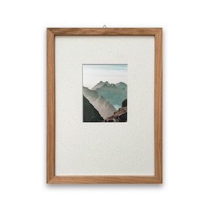 May include: A framed landscape photograph of a mountain range. The image is set within a light brown wooden frame with a white mount. The mountain range is shown in shades of green and blue.