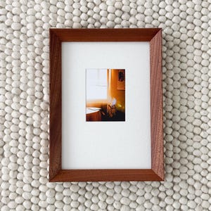 May include: A framed photograph with a warm, inviting interior scene. The frame is a rich, dark brown, and the photograph features a window with sunlight streaming in. The photograph is set against a textured, off-white background.