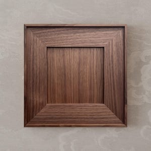 Pode incluir: Moldura quadrada de madeira com um centro rebaixado. A moldura é feita de madeira marrom escura com grão visível, e o design apresenta uma borda chanfrada. O fundo é cinza claro com um padrão sutil.