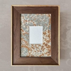 Op de afbeelding: Een rechthoekige houten fotolijst met een gouden binnenrand. De lijst bevat een gemarmerd papierontwerp in tinten grijs, wit en bruin. Een wit vierkant is gecentreerd in het frame, waardoor een gelaagd effect ontstaat.