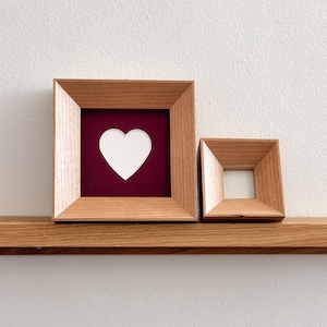 May include: Two wooden picture frames. One frame contains a white heart on a burgundy background. The other frame is empty. Both frames are square and sit on a wooden shelf against a white wall.