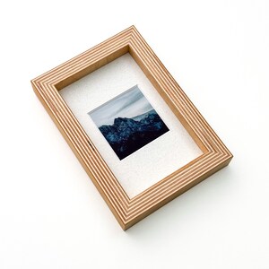 May include: A rectangular wooden picture frame with a landscape photograph. The frame is made of light-colored wood with a layered design. The photograph features a mountain range under a cloudy sky. The frame is angled on a white surface.