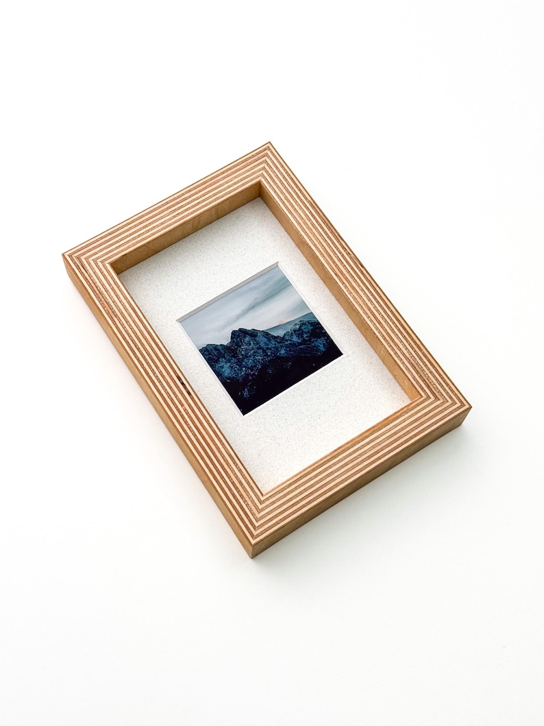 May include: A rectangular wooden picture frame with a landscape photograph. The frame is made of light-colored wood with a layered design. The photograph features a mountain range under a cloudy sky. The frame is angled on a white surface.