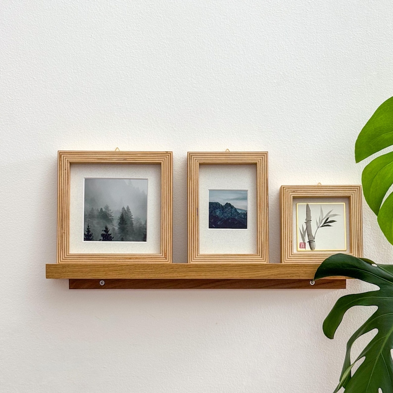 May include: Three framed pictures with light wood frames are displayed on a wooden shelf. The pictures feature a forest scene, a mountain landscape, and a bamboo illustration. The shelf is mounted on a white wall.