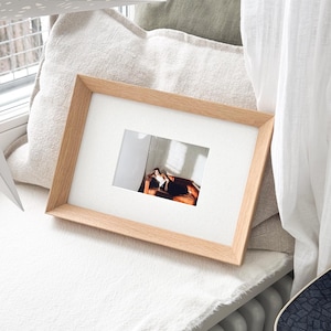 May include: A rectangular wooden picture frame with a white mat, displaying a photograph. The frame is light brown and rests on a white surface, near a window. The photograph shows a person on a brown couch.
