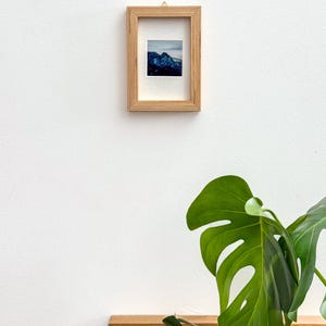 May include: A small framed print featuring a mountain landscape with blue and white tones. The frame is made of light-colored wood and has a textured design. The print is mounted on a white mat, and the frame is hanging on a white wall.