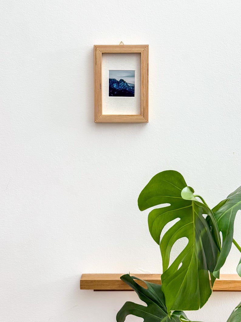 May include: A small framed print featuring a mountain landscape with blue and white tones. The frame is made of light-colored wood and has a textured design. The print is mounted on a white mat, and the frame is hanging on a white wall.