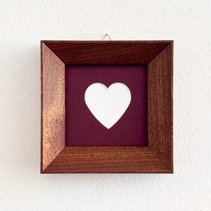 May include: Square wooden frame with a dark brown finish. The frame holds a white heart shape against a burgundy background. The frame is hanging on a white wall.