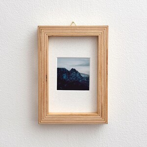 May include: A framed landscape photograph featuring a mountain range under a cloudy sky. The photograph is set within a light brown wooden frame with a ridged design, mounted on a white wall.