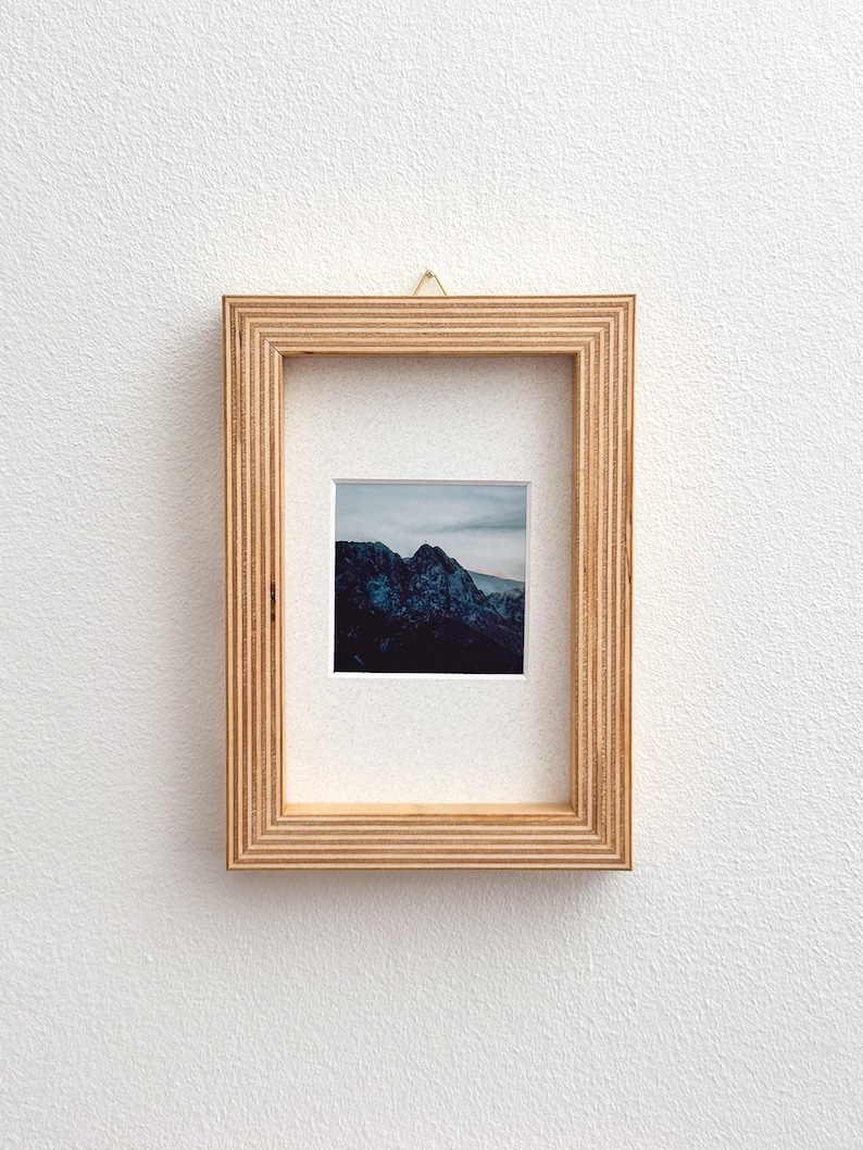 May include: A framed landscape photograph featuring a mountain range under a cloudy sky. The photograph is set within a light brown wooden frame with a ridged design, mounted on a white wall.