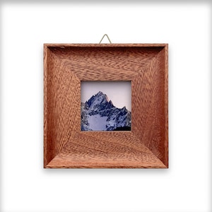 Puede incluir: Marco de fotos cuadrado de madera con una impresión de paisaje de montaña. El marco es de madera marrón rojizo y tiene un pequeño gancho de latón para colgar. La impresión presenta un pico de montaña cubierto de nieve.