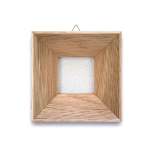 May include: Square wooden picture frame with a natural wood grain finish. The frame has a recessed square opening and a small metal hanger at the top for wall mounting. The frame is empty, with a white backing visible.