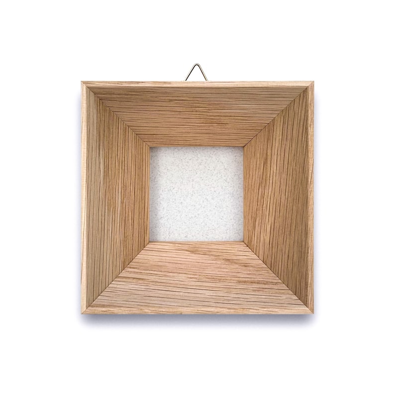 May include: Square wooden picture frame with a natural wood grain finish. The frame has a recessed square opening and a small metal hanger at the top for wall mounting. The frame is empty, with a white backing visible.