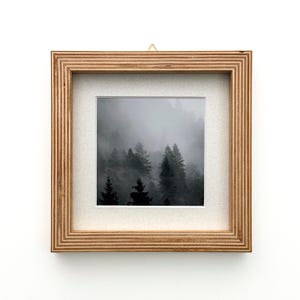 May include: Square framed art print featuring a black and white photograph of a misty forest. The artwork is encased in a natural wood frame with a white mat, and a small gold-colored hook for hanging.
