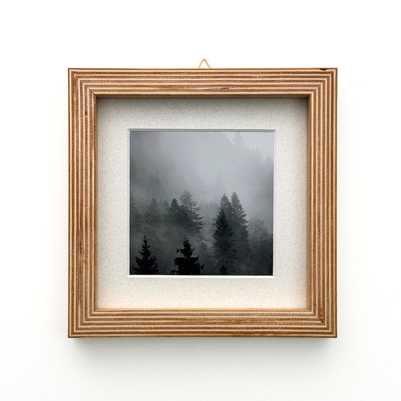 May include: Square framed art print featuring a black and white photograph of a misty forest. The artwork is encased in a natural wood frame with a white mat, and a small gold-colored hook for hanging.