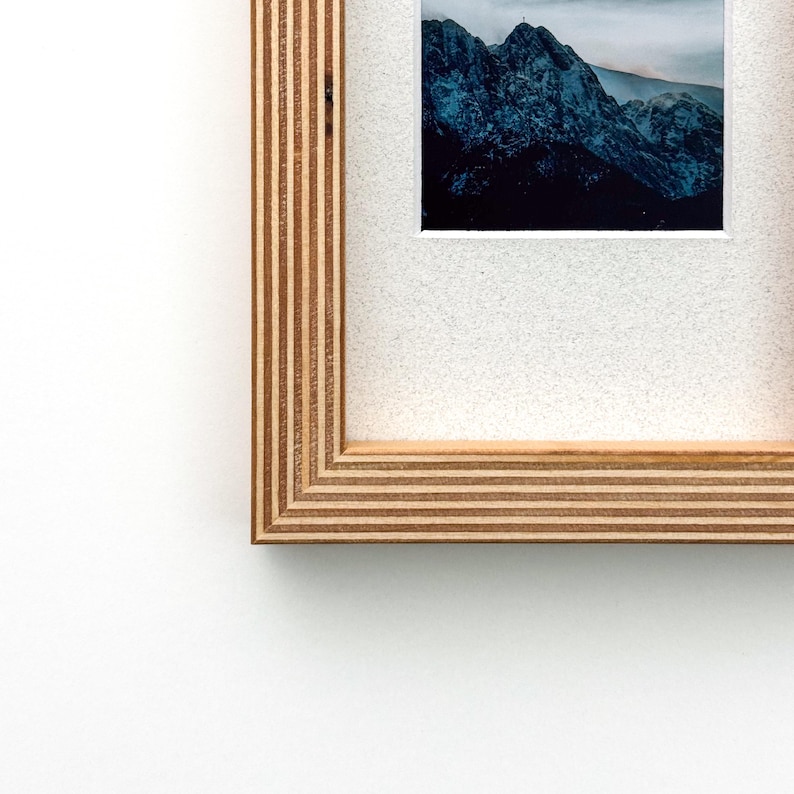 May include: A framed photograph featuring a mountain landscape with a dark blue and gray color palette. The frame is made of light brown wood with a layered design. The photograph is mounted on a white background.