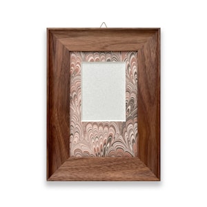 May include: A rectangular wooden picture frame with a warm brown finish. The frame has a marbled paper backing in shades of brown, pink, and white. The center has a white matting around the picture space. A small metal hanger is attached to the top.