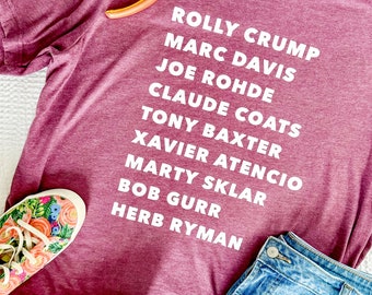 May include: A maroon t-shirt with white text listing the names "Rolly Crump, Marc Davis, Joe Rohde, Claude Coats, Tony Baxter, Xavier Atencio, Marty Sklar, Bob Gurr, Herb Ryman".