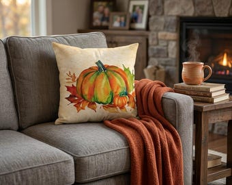 May include: A square throw pillow featuring a watercolor-style illustration of a large orange and green pumpkin surrounded by autumn leaves on a cream-colored background. The pillow is placed on a gray sofa next to a stack of books and a mug.
