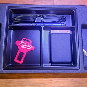 May include: A black plastic center console organizer tray with multiple compartments. It contains a pair of eyeglasses, a pen, a pink Milwaukee branded seatbelt buckle cover, a rectangular power bank, a small metal cylinder, and a silver house key.
