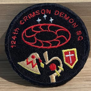 Op de afbeelding: Een ronde, zwarte geborduurde patch met de tekst "124th CRIMSON DEMON SQ" in het rood. De patch heeft rode en gele grafische ontwerpen, waaronder een kruis in een gele schild.