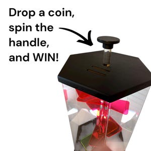 Puede incluir: Una m&aacute;quina de premios hexagonal transparente con una tapa negra y un asa. La m&aacute;quina contiene formas rojas y verdes. El texto de la izquierda dice "Drop a coin, spin the handle, and WIN!"