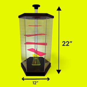 Puede incluir: Una vitrina hexagonal transparente con base y parte superior negras, con estantes rojos. La vitrina mide 56 cm de alto y 30 cm de ancho, con un poste central y una perilla negra en la parte superior. El fondo es amarillo brillante.