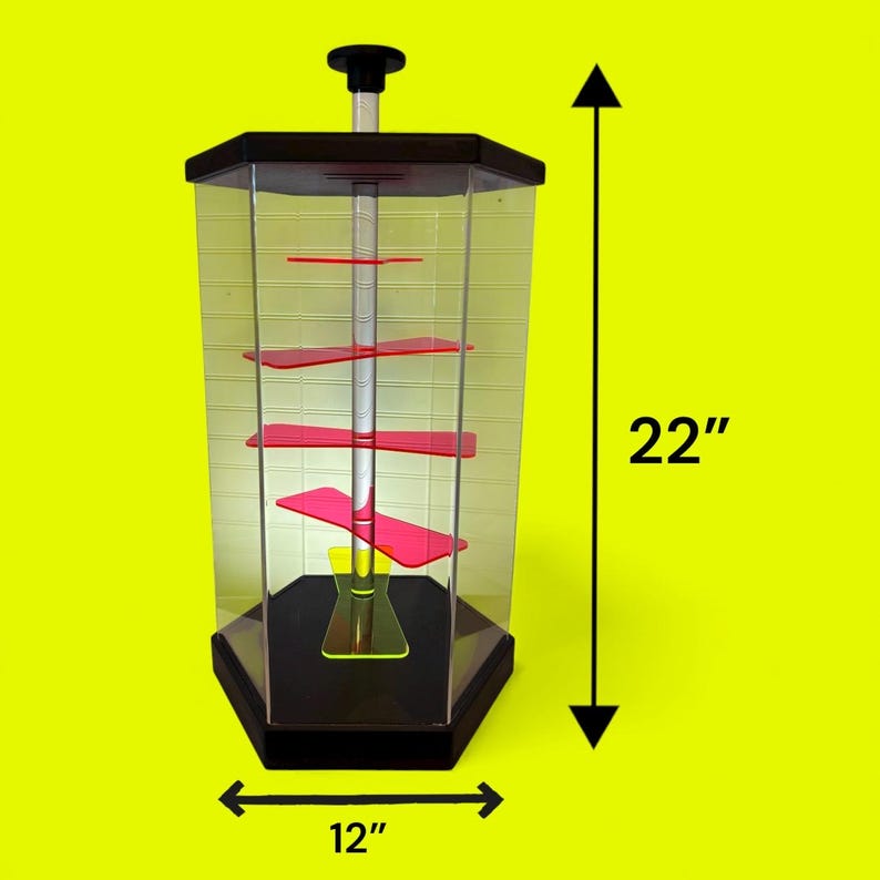Puede incluir: Una vitrina hexagonal transparente con base y parte superior negras, con estantes rojos. La vitrina mide 56 cm de alto y 30 cm de ancho, con un poste central y una perilla negra en la parte superior. El fondo es amarillo brillante.